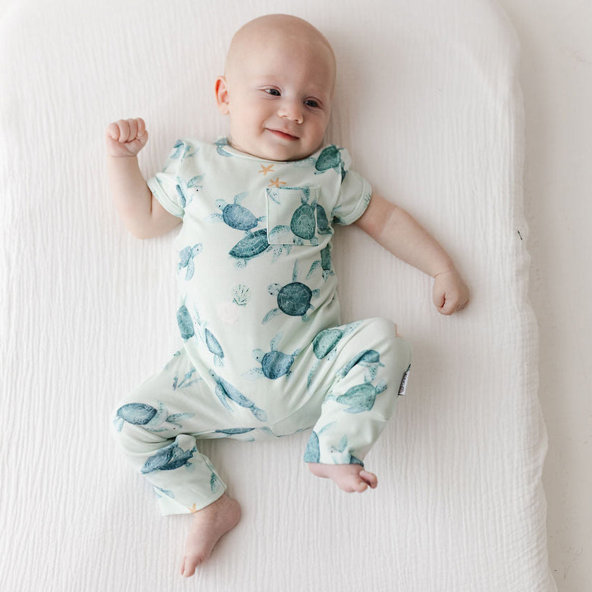 A baby lying on a white muslin changing pad, wearing a short-sleeve footless onesie with a light green and teal sea turtle print.