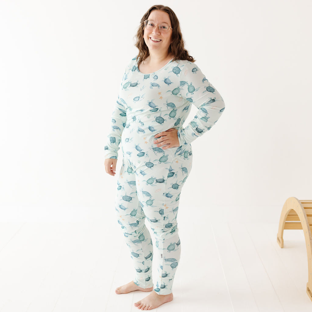 A woman wearing a long-sleeve, one-piece pajama set with a blue and green leaf pattern on a white background, standing in a bright studio setting.
