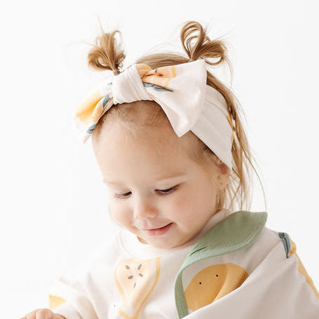 A baby sitting on a white surface wearing a cream-colored wrap blanket with green and yellow cartoon animal prints and a matching white bow headband.