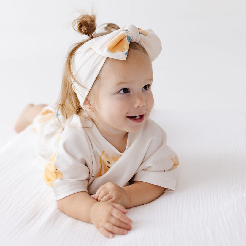 A baby lying on their stomach on a white textured blanket, wearing a cream-colored short-sleeve romper with a yellow floral print and a matching cream headband with a bow.