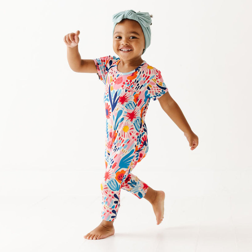 A smiling toddler walking barefoot on a white studio background, wearing a short-sleeve one-piece pajama with a colorful tropical floral print and a light blue knotted headband.