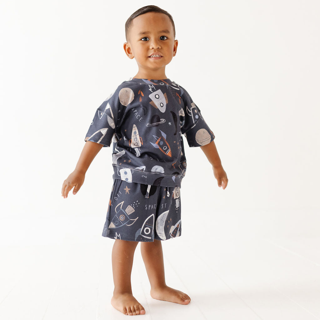 A toddler boy wearing a matching short-sleeve t-shirt and shorts pajama set with a space-themed print of rockets, planets, and stars on a dark navy blue background, standing against a plain white studio background.