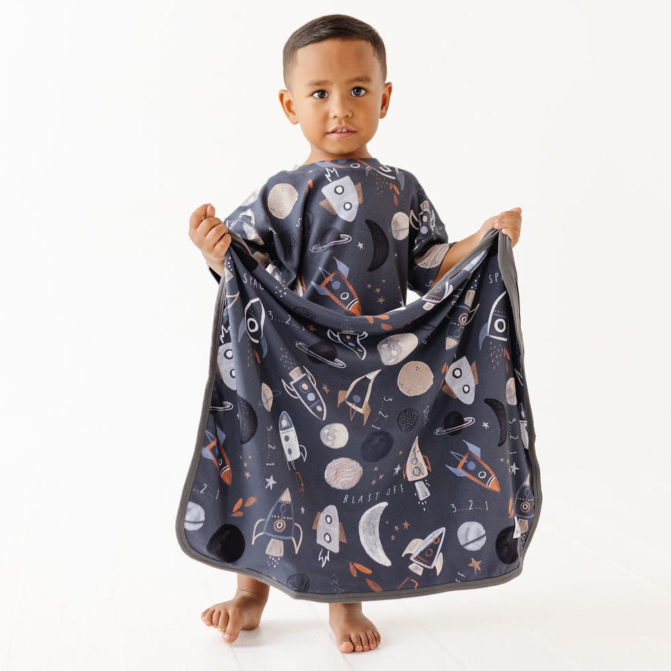 A young child standing barefoot against a plain white background, holding up a dark navy blanket or poncho with a space-themed print of rockets, planets, moons, and stars.