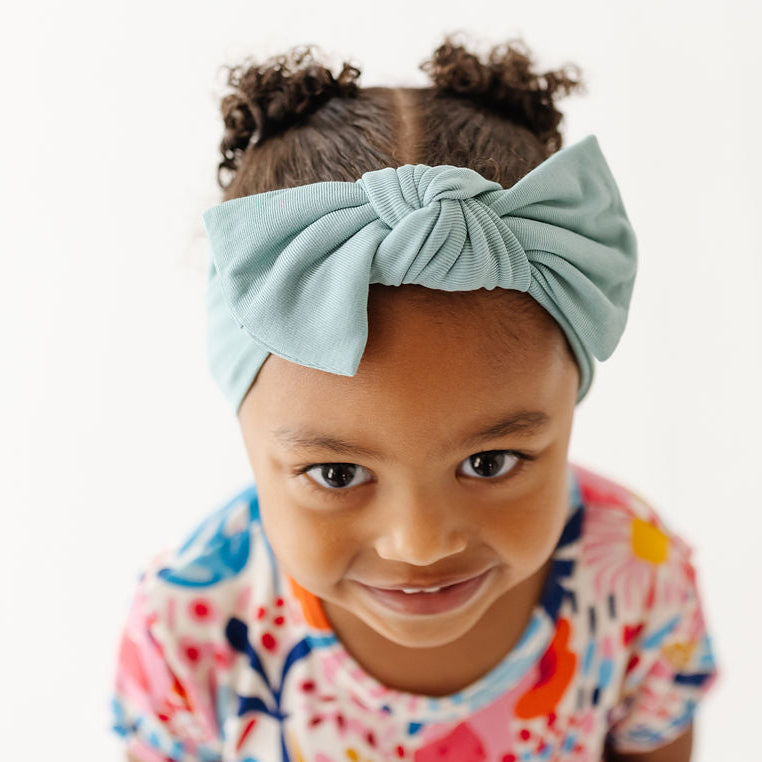 A young child with two small puffs of hair wearing a light blue knotted fabric headband and a colorful floral-patterned t-shirt, photographed against a clean white background.