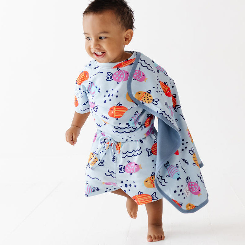 A smiling toddler walking barefoot on a white studio floor while wearing a light blue wrap dress with an orange, pink, and navy ocean-fish print. A matching light blue towel is draped over one shoulder.