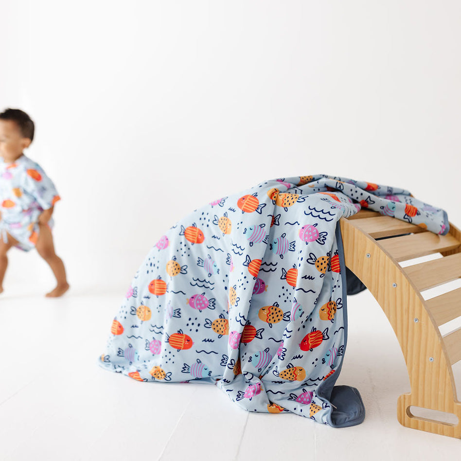 A baby walking outfit with a matching light blue patterned blanket draped over a wooden bench in a bright studio. The outfit features a playful multicolor pattern of fish, sea plants, and sea life on a light blue background.
