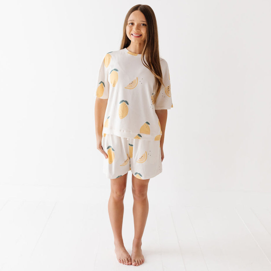 A woman modeling a matching short-sleeve pajama set with a lemon print, consisting of a cream-colored top and shorts, against a clean white studio background.