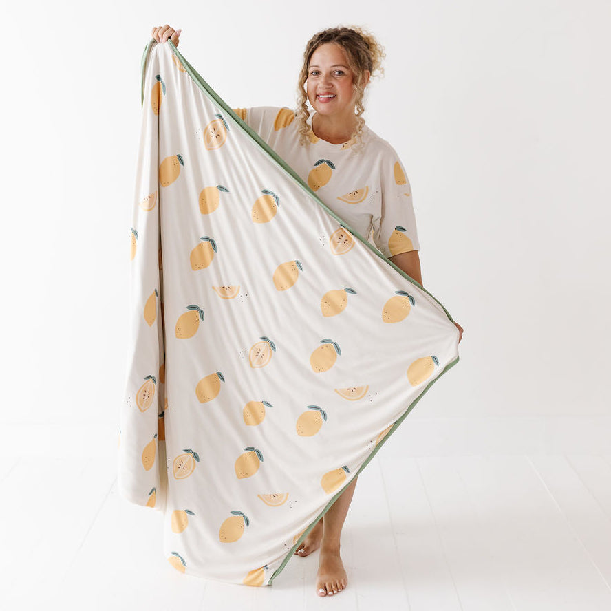 A woman stands barefoot against a white studio background, holding up a large cream-colored blanket or sheet with a repeating yellow-orange citrus fruit print and a green border.