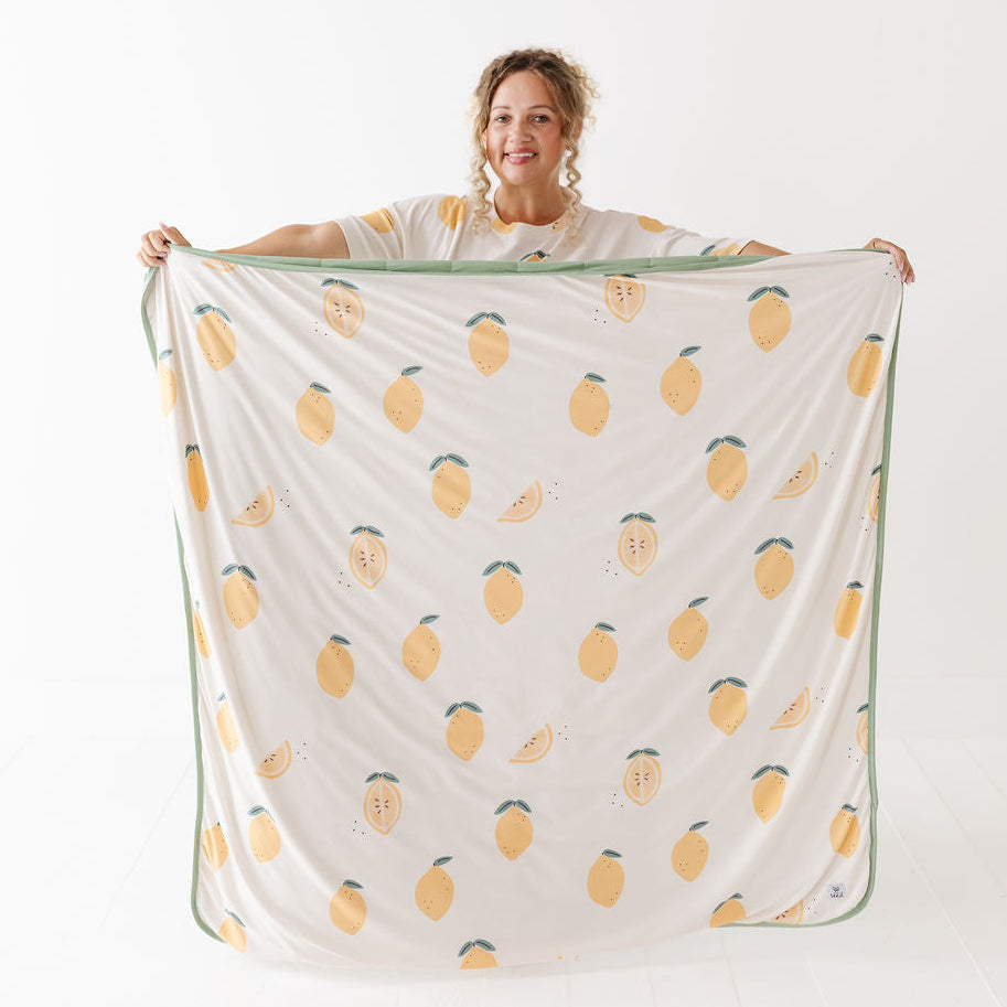 A woman holds up a large blanket or throw with a repeating yellow lemon print and a green border, photographed against a clean white studio background.