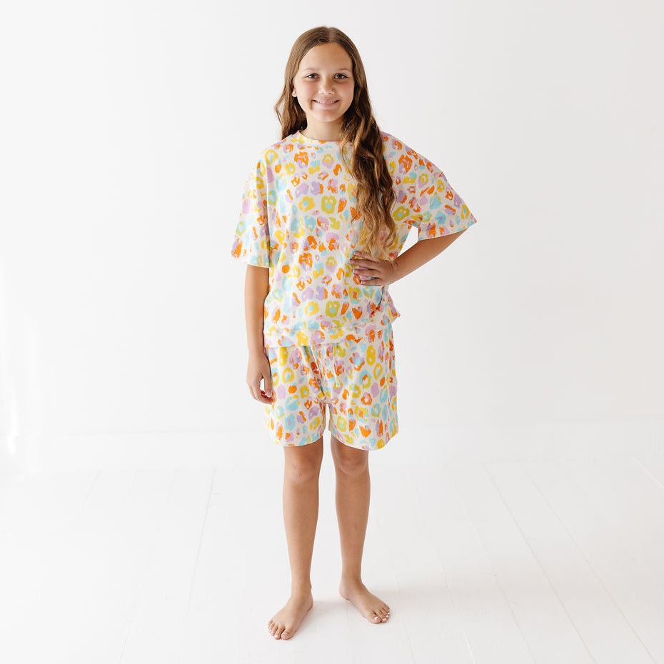 A young girl stands barefoot in a bright white studio, wearing a short-sleeve medical-style gown and matching shorts set with a colorful cartoon pattern of characters and medical icons on a white background.