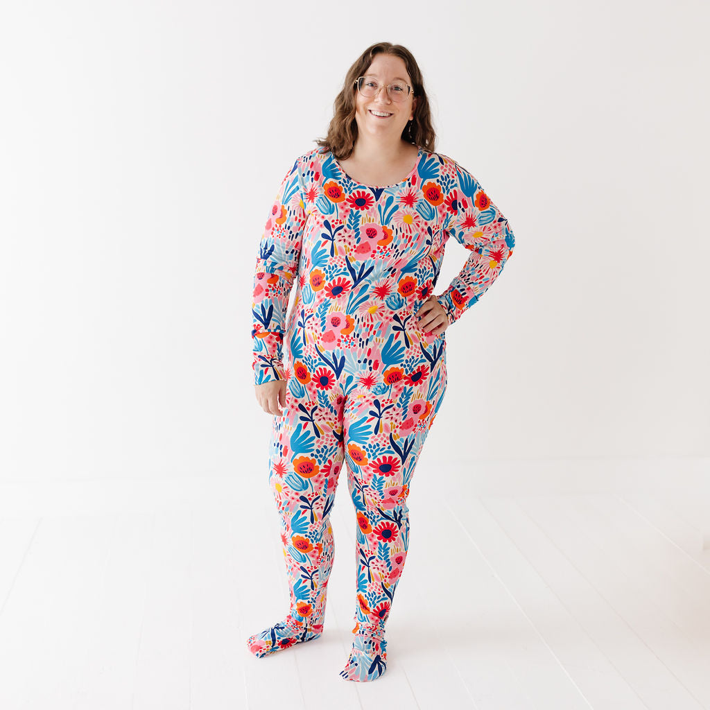 A woman modeling a long-sleeve, full-body footed pajama set with a vibrant floral print in red, orange, blue, and white, standing against a plain white studio background.