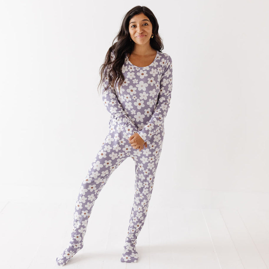 A woman modeling a one-piece footed pajama set with a white daisy floral print on a light lavender-gray background, standing against a clean white studio backdrop.