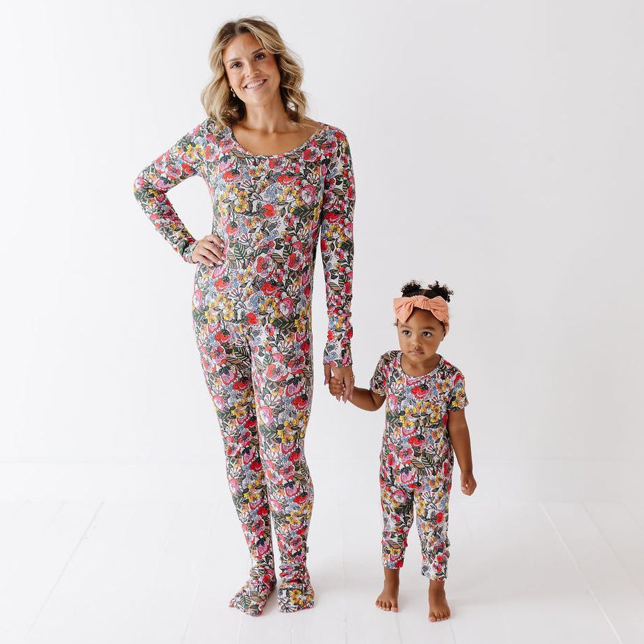 A woman and a young child standing hand in hand, both wearing matching long-sleeve pajama sets with a dense multicolor floral print, against a clean white studio background.