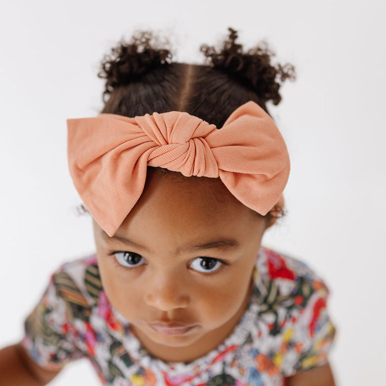 A toddler girl looking up at the camera, wearing a large peach-colored fabric bow headband and a colorful floral print t-shirt against a plain white background.