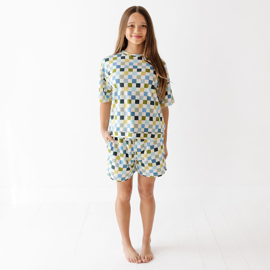A young girl stands barefoot against a bright white studio background, wearing a matching short-sleeve top and shorts set with a blue, yellow, and white geometric checkerboard pattern.