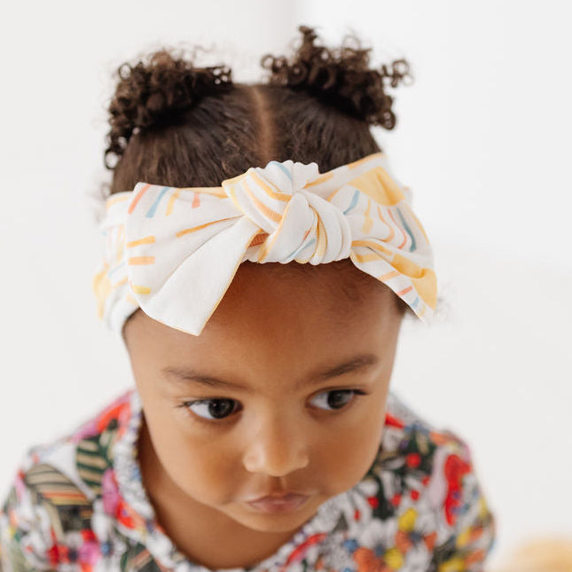 A toddler girl with her hair in two buns wears a white fabric bow headband with yellow and blue stripes, paired with a colorful floral t-shirt, sitting on a wooden bench against a white background.