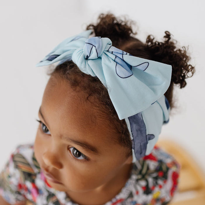 A toddler girl with curly dark hair wearing a light blue fabric bow headband with a dark blue abstract pattern, paired with a colorful floral dress, sitting on a wooden slatted bench against a white wall.