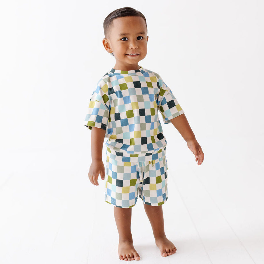 A young child standing barefoot against a clean white studio background, wearing a matching short-sleeve top and shorts set with a multicolor geometric check pattern.
