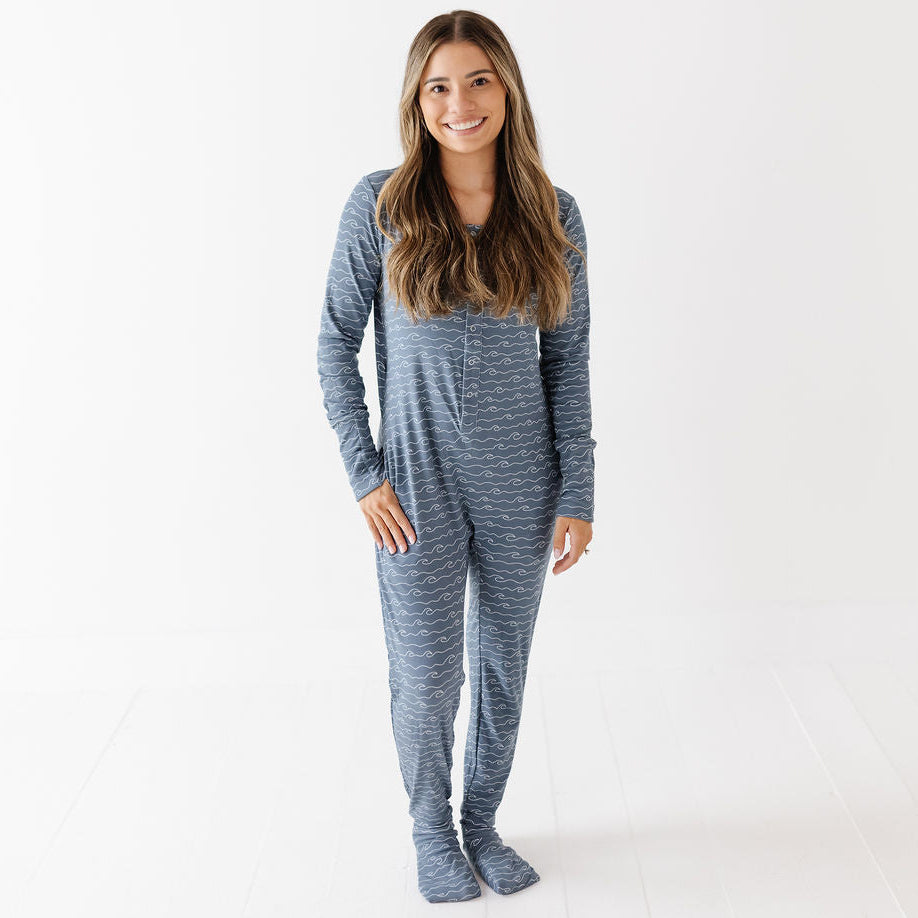 A woman modeling a long-sleeve, one-piece footed pajama set with a heathered blue and gray pattern, standing against a clean white studio background.