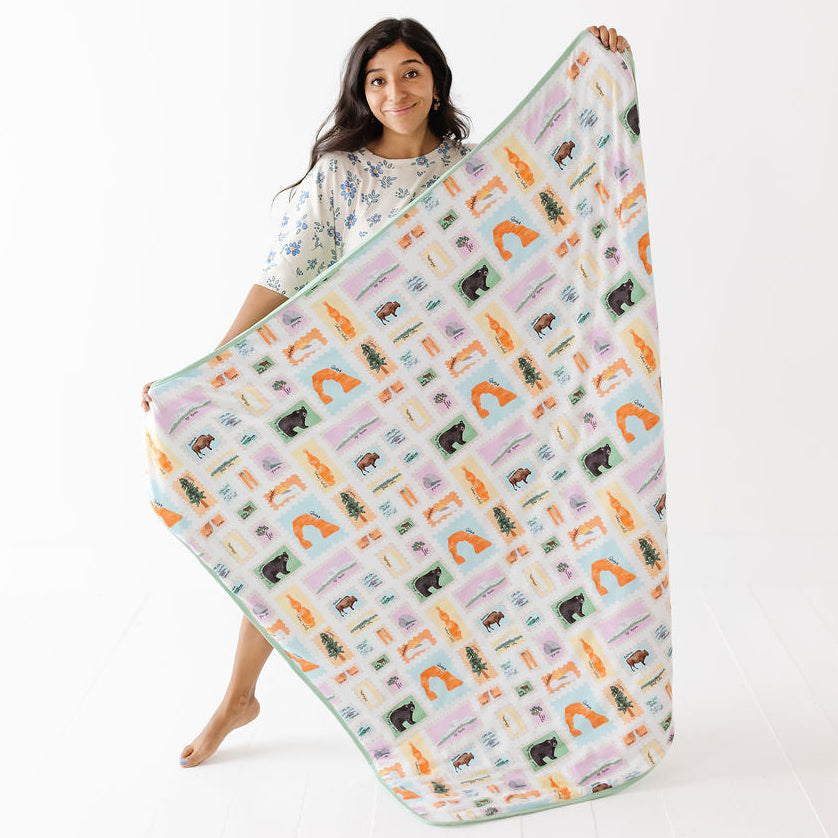 A woman holds up a large fleece blanket with a colorful forest pattern featuring trees, animals, and arches on a white background.