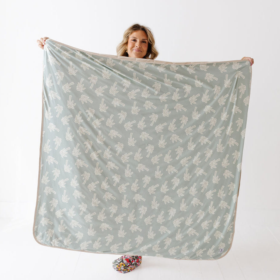 A woman holds up a large square blanket or throw with a sage green background and a repeating white leaf pattern, shown against a plain white studio background.