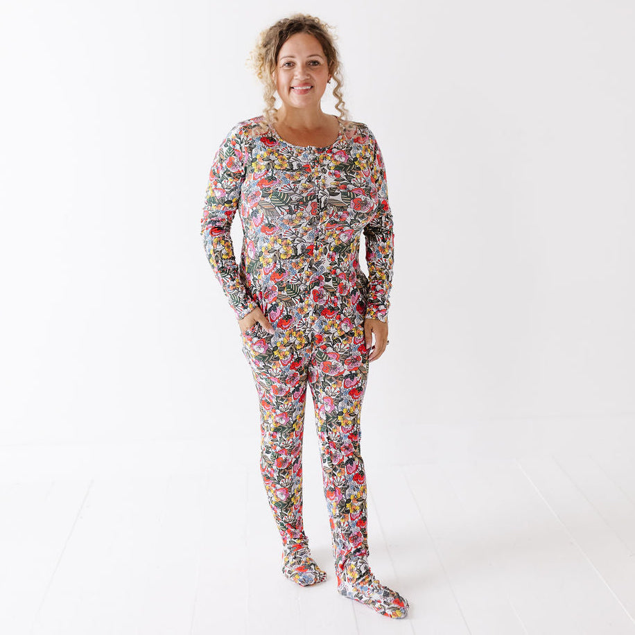A woman modeling a long-sleeve footed pajama set with a dense multicolor floral and abstract print, standing against a plain white studio background.