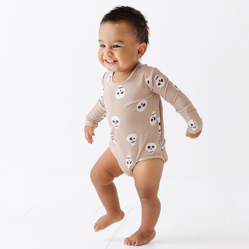 A smiling toddler walking barefoot on a clean white studio floor, wearing a long-sleeve beige bodysuit covered in small white skulls with black outlines.