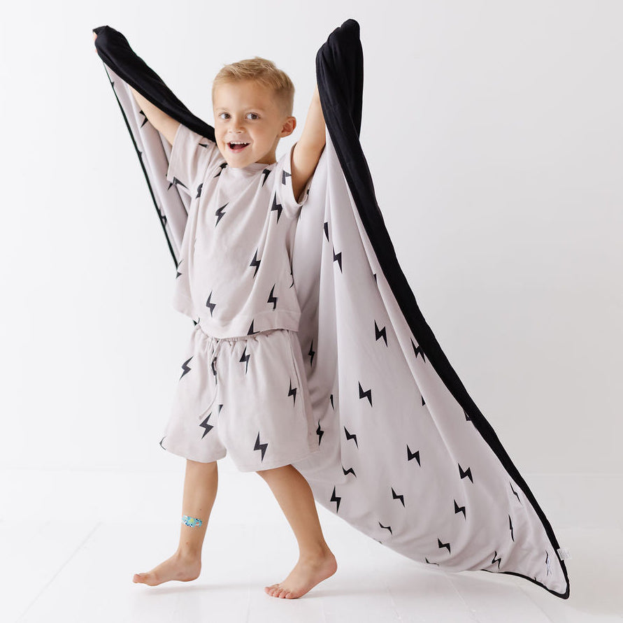 A young child wearing a matching light gray pajama set with a black lightning bolt pattern, playfully holding a large wing-shaped blanket with black trim outstretched behind them against a clean white studio background.