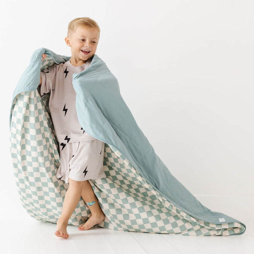 A young child stands barefoot on a white studio floor, wrapped in a large duvet or blanket with a pale blue top section and a mint green and white checkerboard patterned underside.