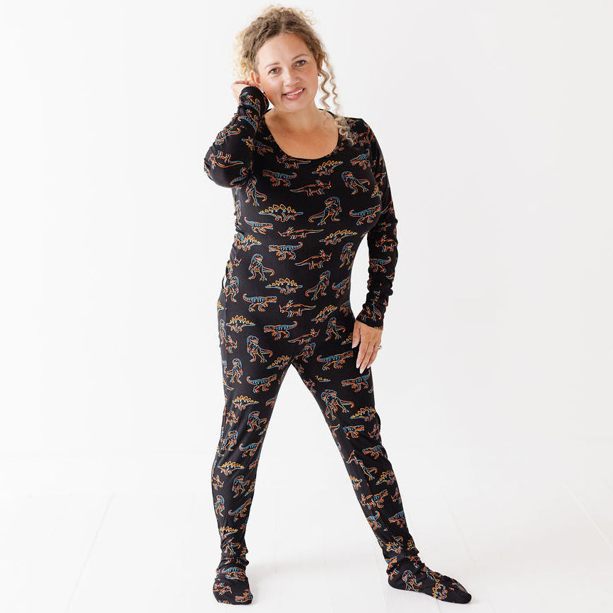 A woman modeling a black long-sleeve footed pajama set with a repeating orange and gold abstract pattern, standing against a plain white studio background.