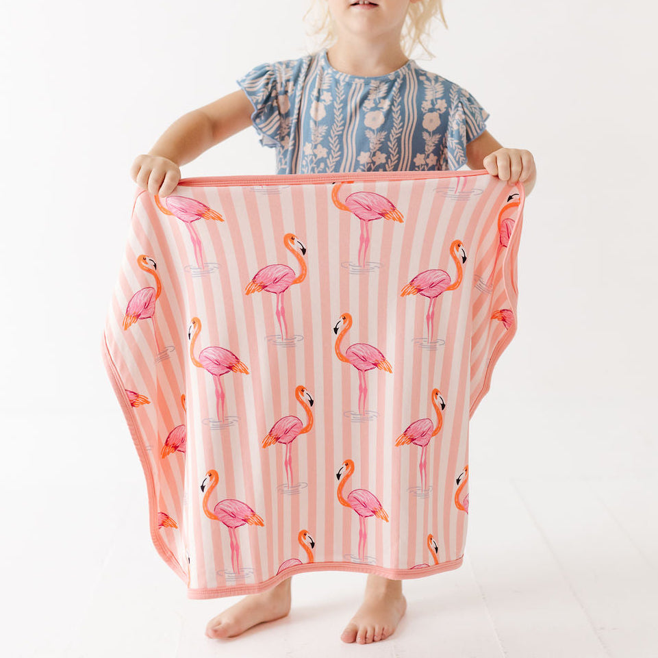 A young child stands barefoot against a white studio background, holding up a large square textile with a pink flamingo print on a white and pink striped base. The child wears a blue patterned top, and the textile appears to be the product being shown.
