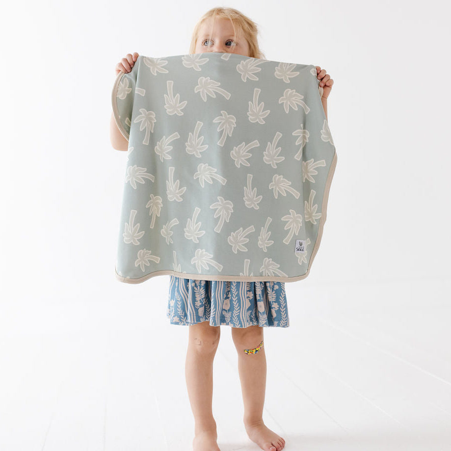 A young child stands barefoot against a white studio background, holding up a square cloth or blanket with a pale sage green base and a repeating cream palm tree pattern. The child is wearing a blue patterned dress.