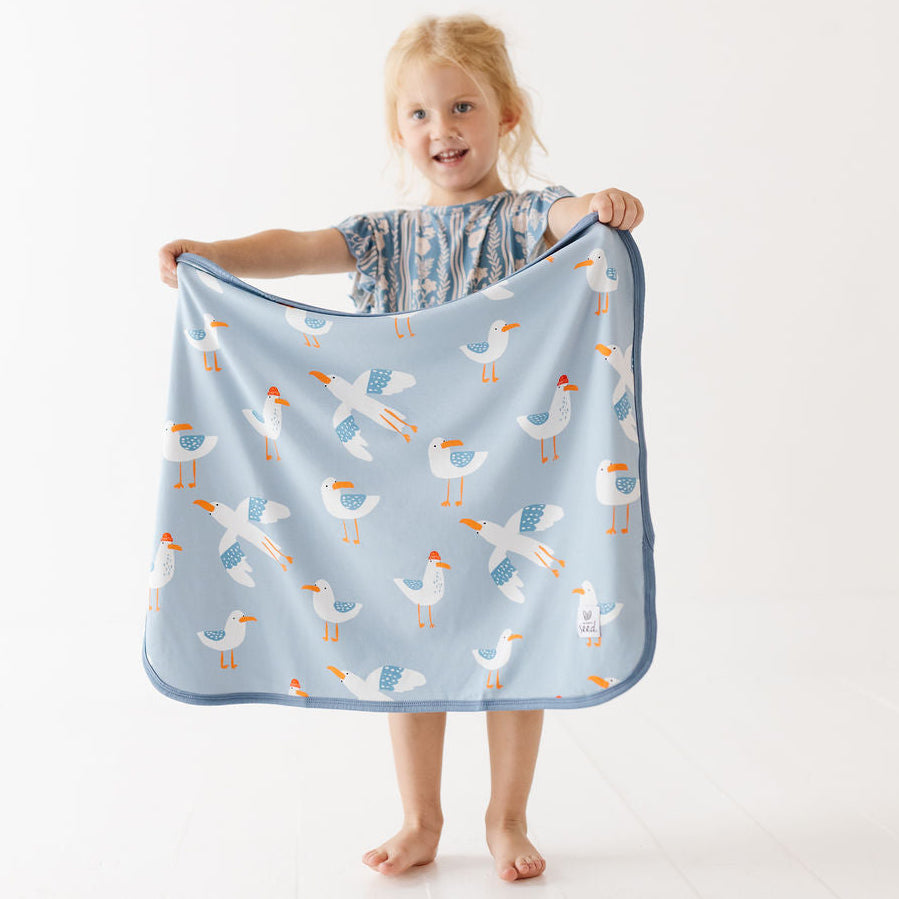 A young blonde girl stands barefoot against a white studio background, holding up a light blue blanket with a playful pattern of white seagulls with orange beaks and blue accents.
