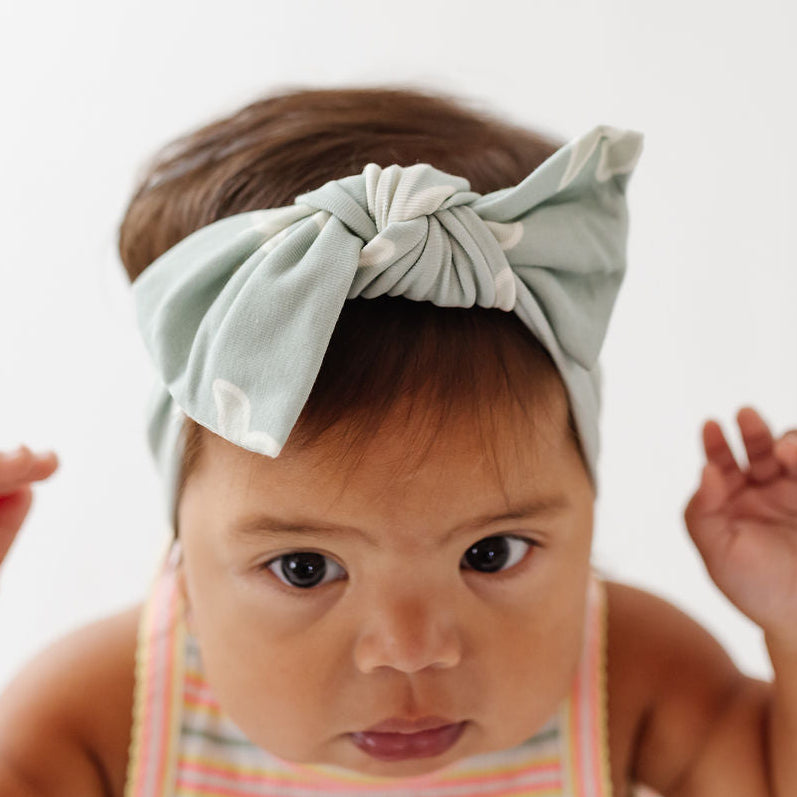 A close-up of a baby wearing a sage green fabric bow headband, looking directly at the camera against a bright white background.