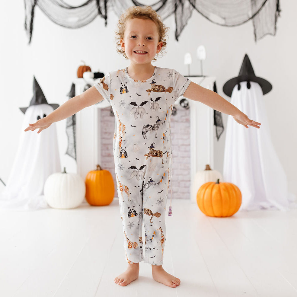 A young child stands barefoot with arms outstretched in a bright white room decorated for Halloween, wearing a short-sleeve pajama top and matching pants with an allover animal print on a light background.