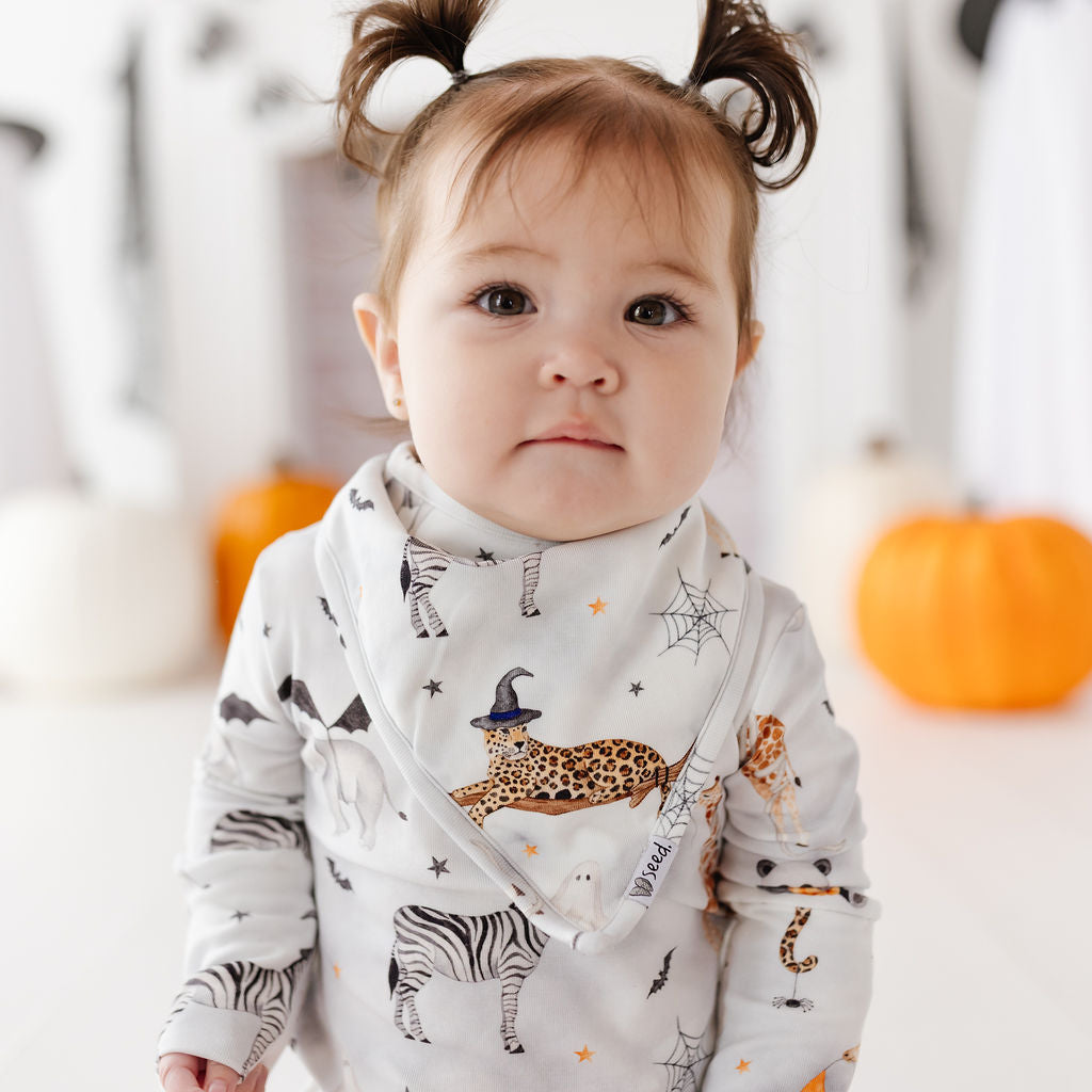 A baby wearing a long-sleeve white bib and matching top with a Halloween animal print, including a leopard, zebra, spiderwebs, and bats. The baby has brown hair styled in two buns and is looking directly at the camera against a softly blurred background with pumpkins.