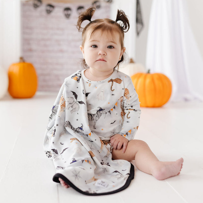A baby sitting on a white floor wearing a Halloween-themed printed blanket or swaddle with bats, ghosts, and pumpkins, posed in a decorated room with pumpkins and ghost props.