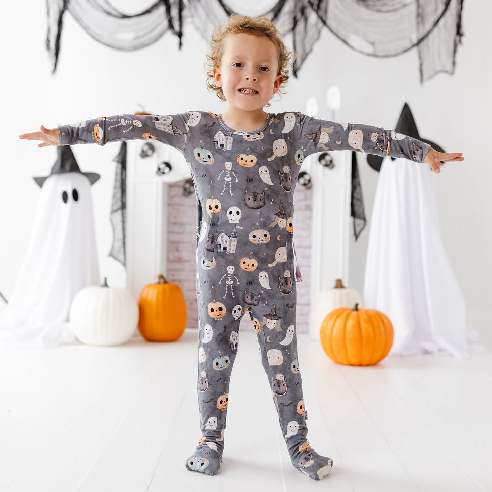 A young child wearing a gray long-sleeve pajama set with a Halloween print of ghosts, pumpkins, and skeletons, standing in a decorated room with white sheets, black witch hats, and pumpkins.