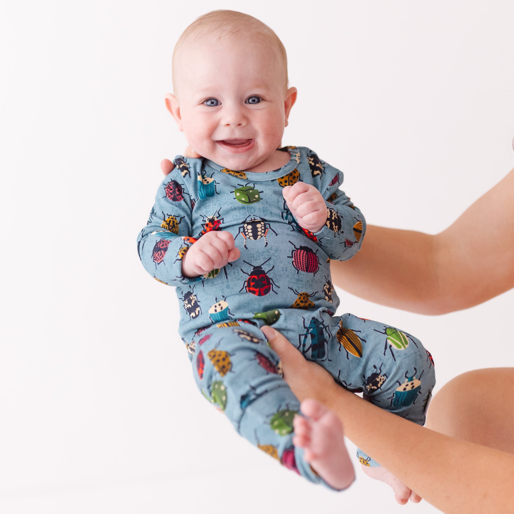Baby wearing a blue onesie with colorful insect patterns, standing next to an adult's arm.