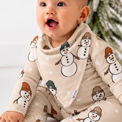 A baby sitting on a white floor wearing a beige footed sleeper with a snowman print, flanked by two small artificial Christmas trees.