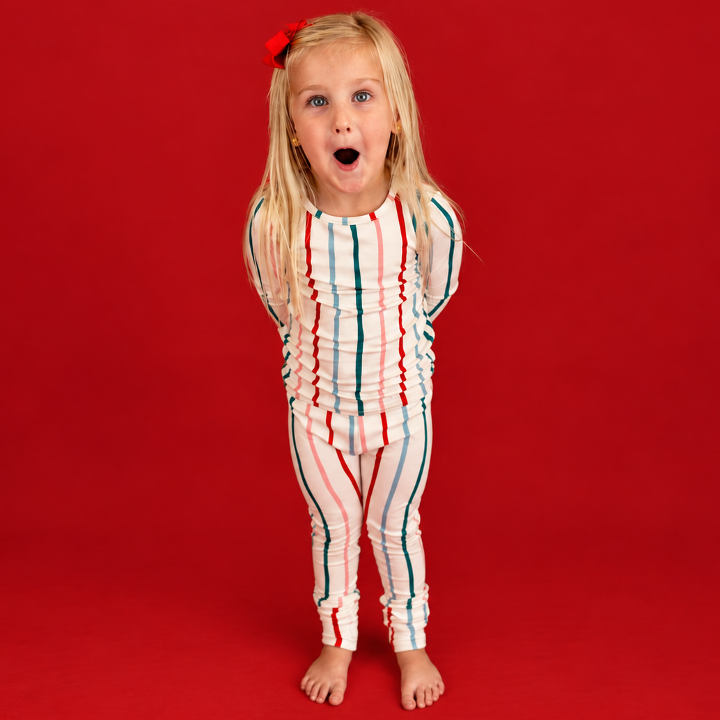 A young blonde girl with a red bow in her hair stands against a solid red studio background, wearing a one-piece long-sleeve swimsuit with vertical red, white, and blue stripes.